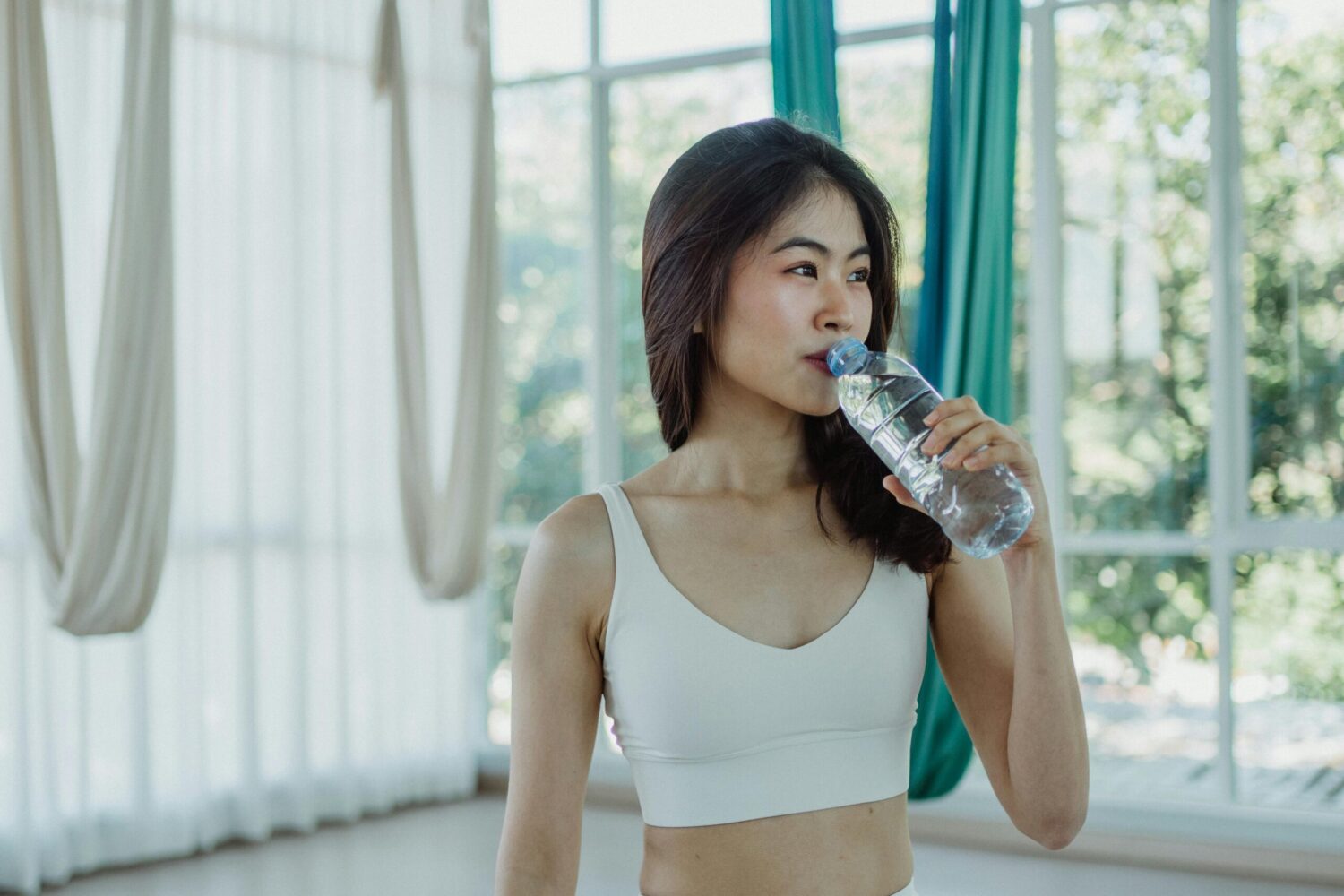 Woman in sportswear drinking water indoors, promoting hydration and wellness.